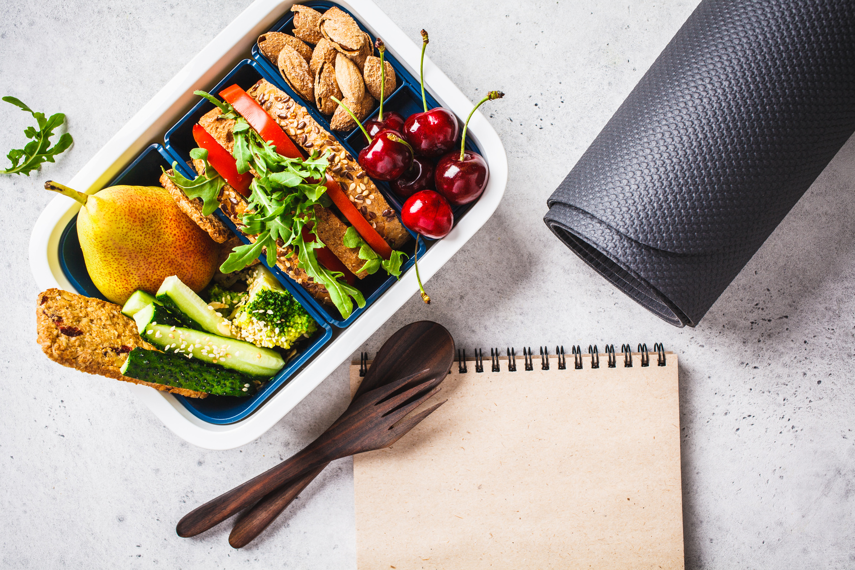 Sport food concept. Lunch box with heathy food, notebook and mat on a gray background.